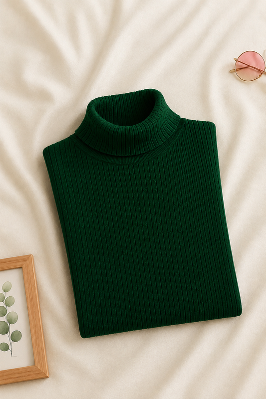 A dark green ribbed turtleneck sweater folded neatly on a cream-colored fabric background, styled with a small picture frame, pink sunglasses, and a green botanical print.