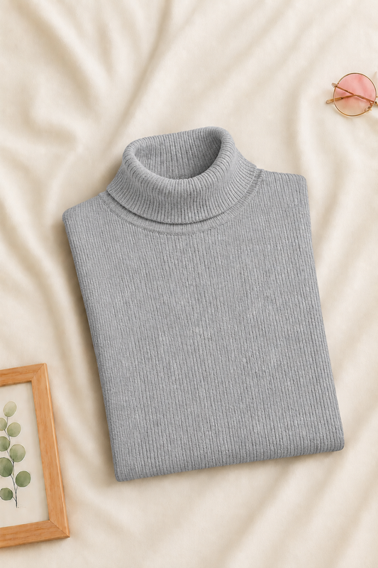 A folded light gray ribbed turtleneck sweater is displayed on a cream-colored fabric background, styled with a small picture frame, pink sunglasses, and a neutral leaf illustration.
